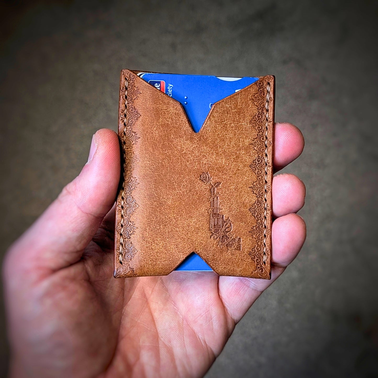 A close-up of a hand holding the El Rey Leather Deadbeat minimalist wallet in cognac, showing the stamped details.