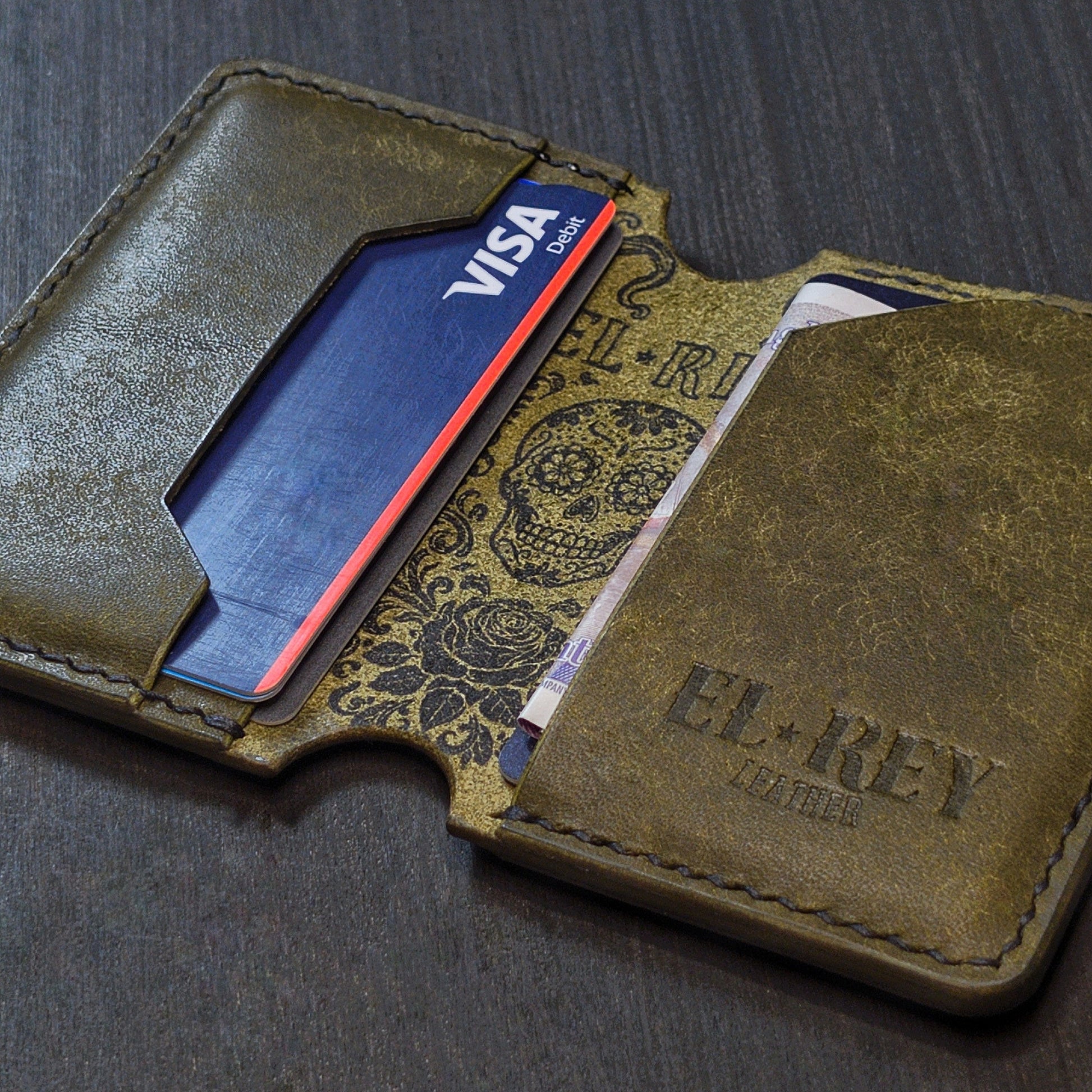 Angled view of El Rey Leather's Lowlife minimalist wallet in olive with cards, showing the ink skull and embossed logo.